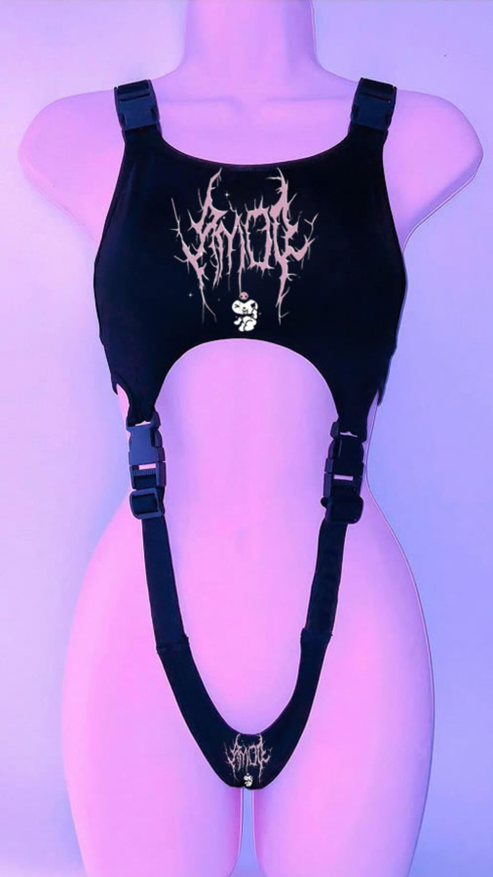 Black harness-style crop top on a mannequin with adjustable buckle straps and side cut-outs. Features a bold white graphic print with skull and stylized text on the chest, plus a smaller matching graphic at the bottom, shown under purple lighting.