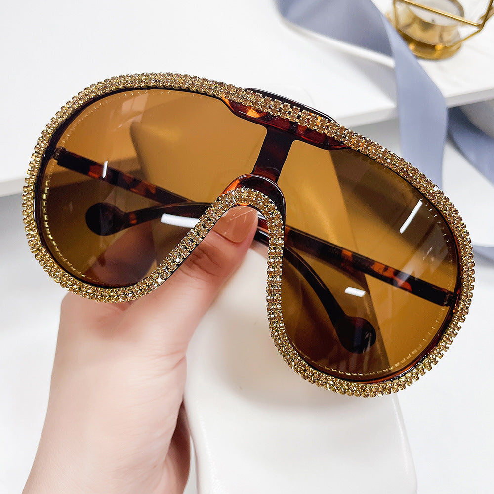 Hand holding oversized amber-lens shield sunglasses with gold rhinestone trim.