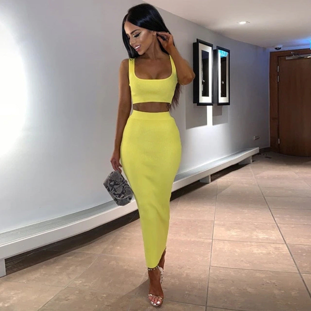 Model in a vibrant chartreuse yellow two-piece outfit featuring a sleeveless square-neck crop top and form-fitting high-waisted midi skirt. Full-body fashion pose in a bright contemporary interior with white walls and tiled flooring, accessorized with a small handbag and strappy heels.