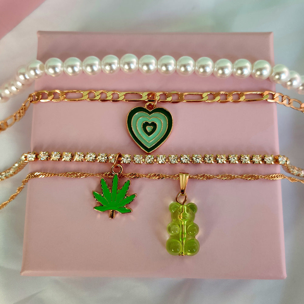 Gold pearl layered necklace set with green heart, leaf & gummy bear pendants – trendy statement jewelry collection on pink and white background.