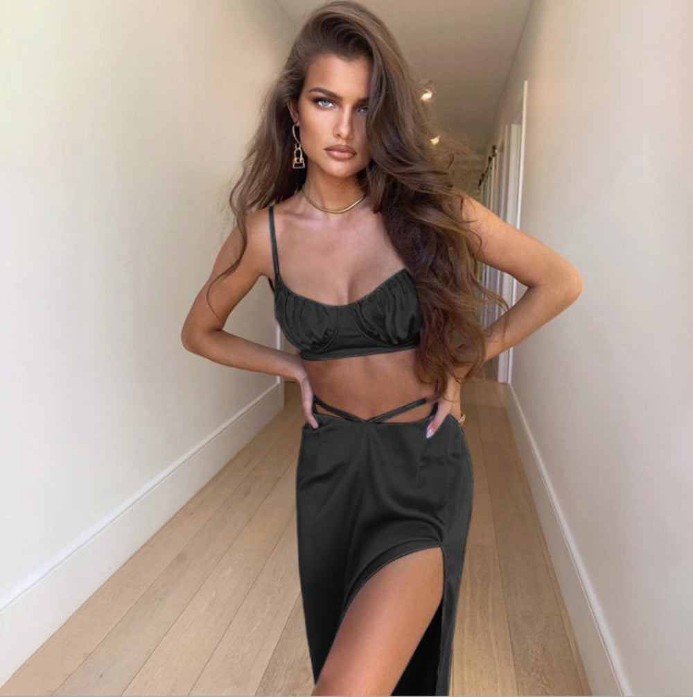 Model in a shiny black matching set: sleeveless ruched crop top with delicate straps and a figure-hugging mini skirt with adjustable side details. Full-body fashion pose with hands on hips in a bright modern hallway, highlighting the glossy fabric and flattering silhouette.