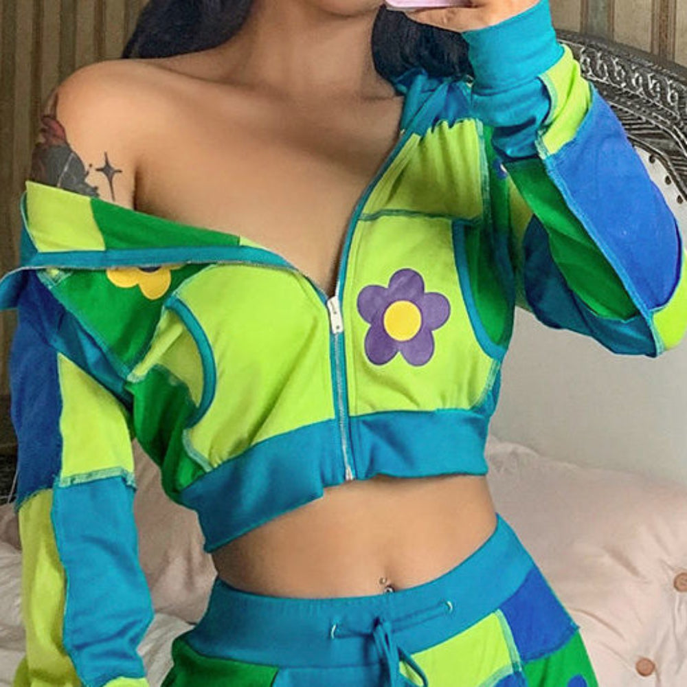 Woman taking a mirror selfie in a vibrant patchwork cropped zip-up hoodie and matching shorts set. The off-shoulder hoodie features lime green, blue, and green color-block panels, daisy flower appliqués, and a partially open front zipper, while the shorts have a drawstring waist and coordinating flower details. Styled with a visible belly button piercing and shoulder tattoo.