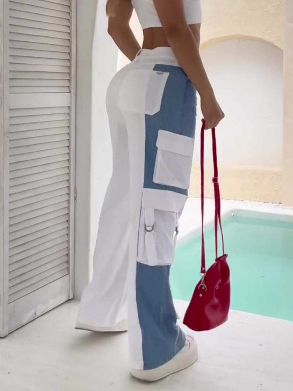 Side and back view of a woman wearing high-waisted color-block cargo jeans featuring white and blue denim panels with multiple large flap pockets and metal hardware. Styled with a white crop top, chunky white sneakers, and carrying a small red crossbody bag, photographed outdoors by a pool.