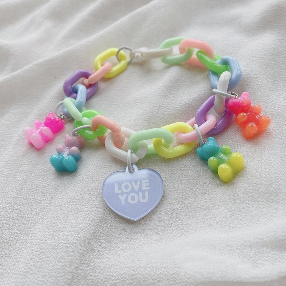 Rainbow pastel gummy bear charm bracelet with a multicolored chain of chunky pastel links in pink, purple, yellow, green, blue and white, featuring colorful translucent gummy bear charms and a light purple heart-shaped pendant engraved with "LOVE YOU", displayed on white fabric.