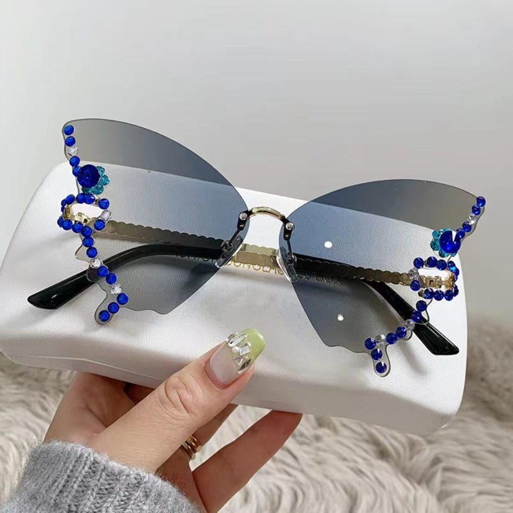 Rimless butterfly sunglasses with gray gradient lenses and blue crystal wing details, held on a white glasses case.