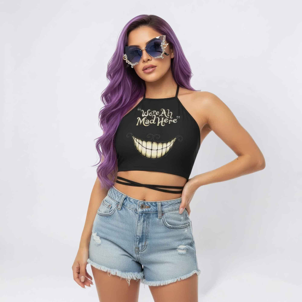 Woman with long vibrant purple wavy hair wearing ornate blue decorative sunglasses and a black halter crop top with the text We're All Mad Here above a large Cheshire Cat grin graphic. The top has a cross-wrap detail at the waist and is paired with light-wash distressed denim shorts.