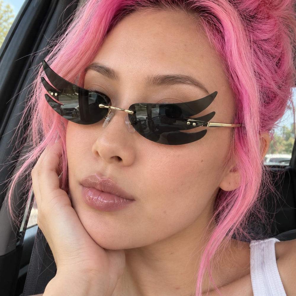 Woman with pink hair wearing black flame-wing sunglasses with gold temples.