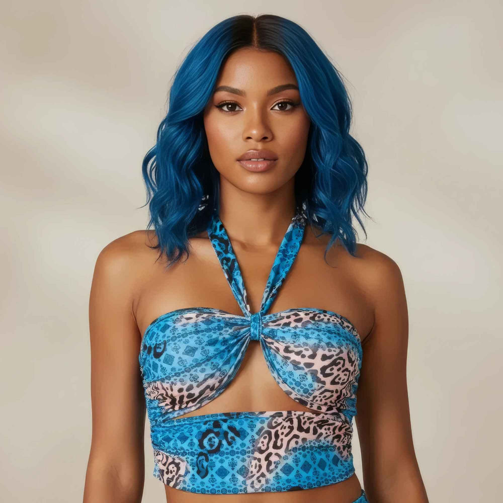 Front portrait of a woman with vibrant blue wavy hair wearing a bold blue patterned halter crop top. The stylish two-piece set features a mix of blue leopard print and geometric patterns with a flattering front twist knot detail and crisscross halter straps.