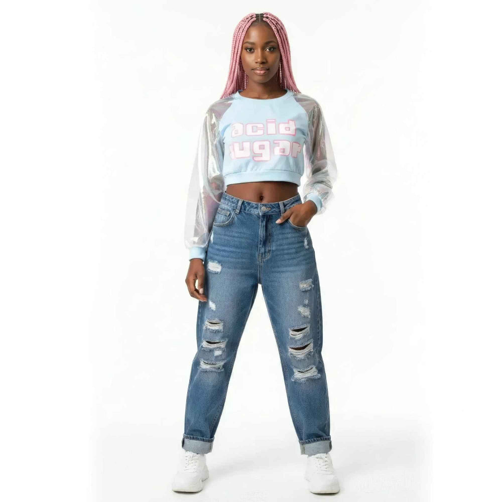Woman with long pink braids modeling a light blue cropped t-shirt featuring bold acid sugar graphic text and sheer iridescent long sleeves. Paired with high-waisted distressed blue denim jeans with multiple rips and white sneakers.