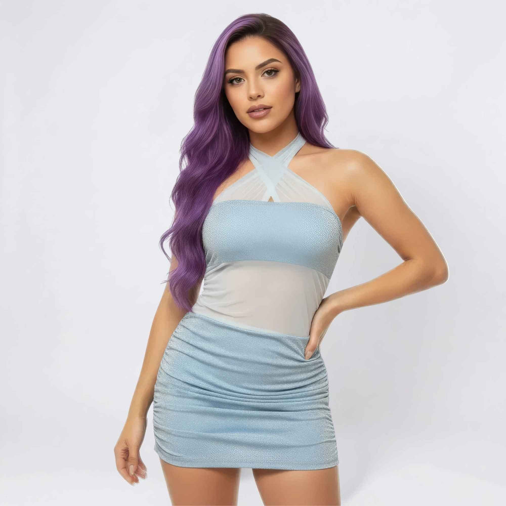 Front view of a woman with long vibrant purple wavy hair modeling a shimmering light blue mini dress. The form-fitting dress features a halter neck with crisscross sheer mesh detail, sparkly fabric, and ruched side detailing on the skirt. She poses confidently with one hand on her hip.
