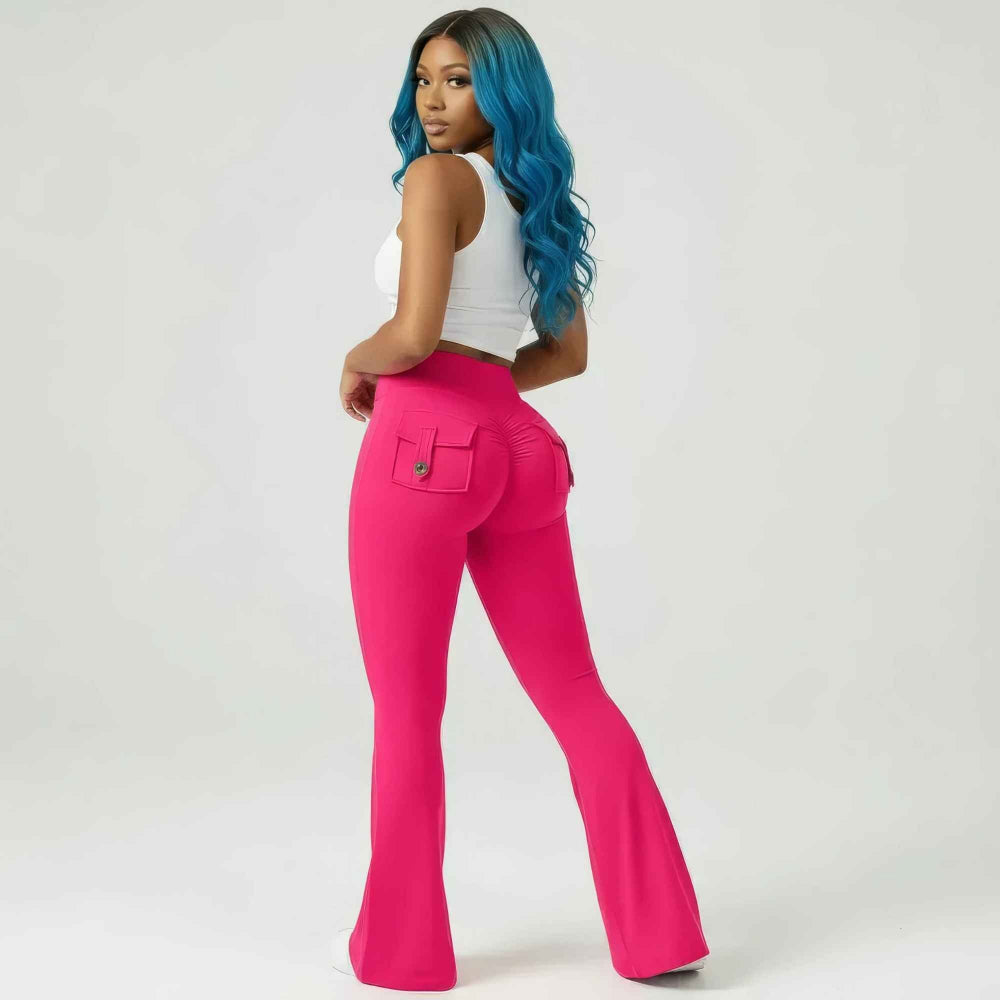 Back three-quarter view of a woman with long vibrant blue wavy hair modeling bright pink high-waisted flared pants with back flap pockets and ruched detailing. She is wearing a white sleeveless cropped tank top, posing confidently while looking over her shoulder to highlight the curve-hugging fit and wide-leg flare.