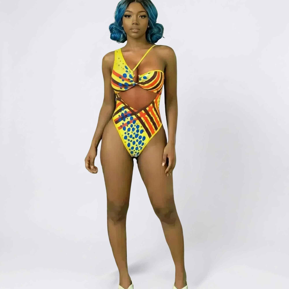 Full-length studio shot of a woman with vibrant blue curly hair modeling a bold yellow one-piece swimsuit with multicolored geometric and polka dot patterns in blue, red, and orange. The swimsuit features a cut-out waist detail, crisscross straps, and a flattering high-cut leg design.