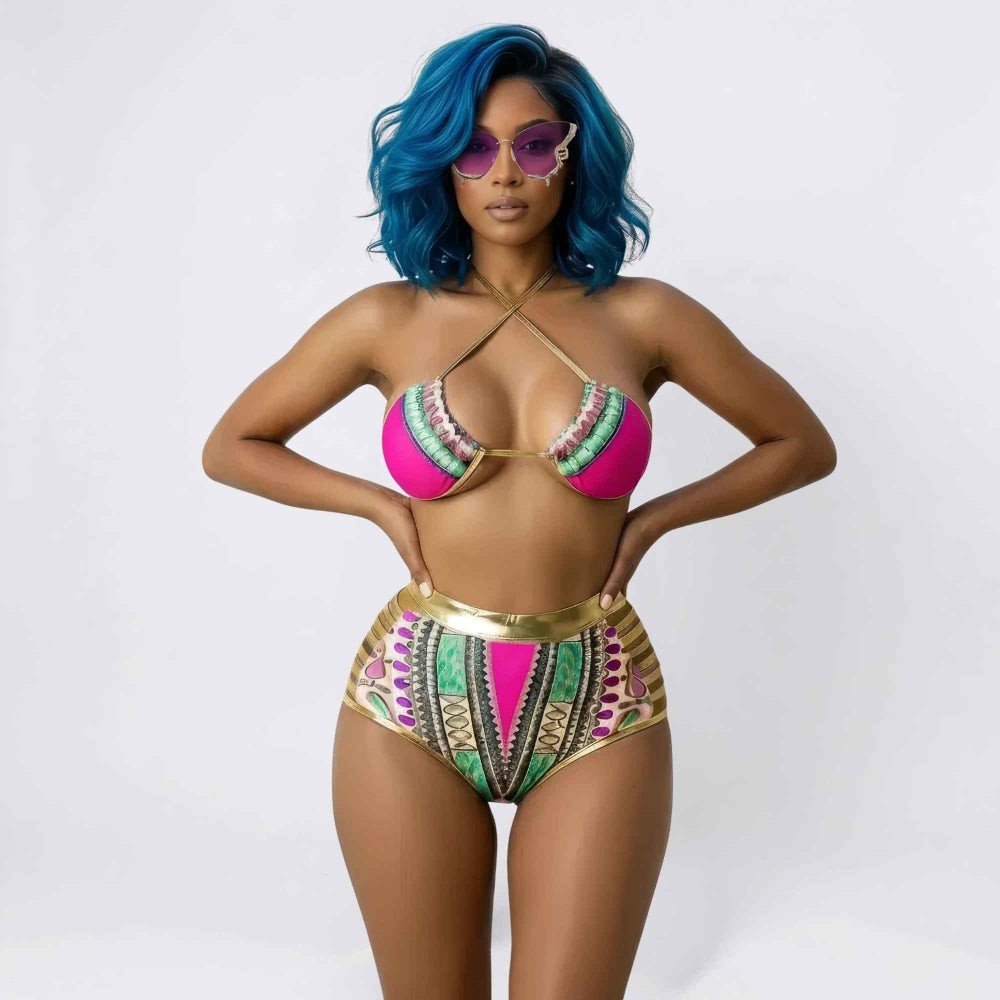 Front view of a confident woman with vibrant blue wavy hair wearing purple decorative sunglasses and a bold two-piece bikini. The set features a bright pink halter triangle top with gold crisscross straps and intricate multicolored patterns, paired with high-waisted bikini bottoms with vibrant tribal designs and a shiny gold waistband