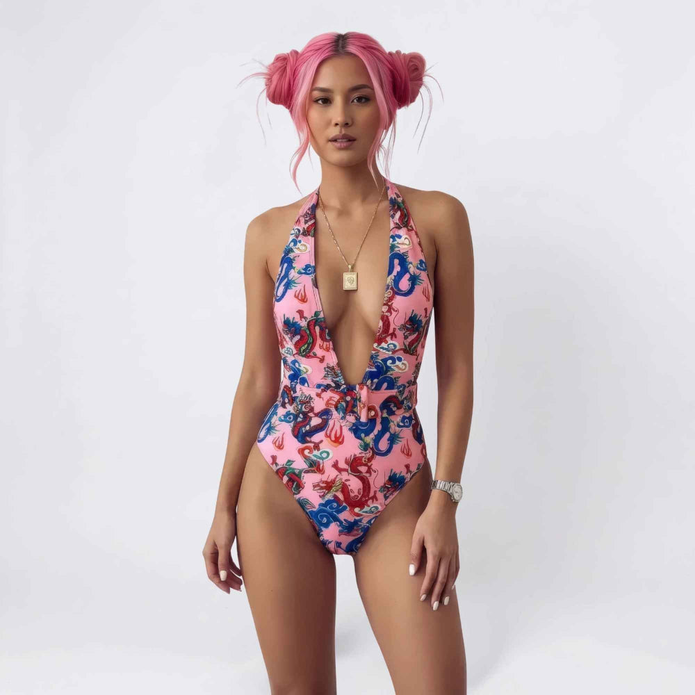 Front view of a woman with bright pink hair in playful space buns modeling a vibrant pink one-piece swimsuit with a deep plunging halter neckline and bold blue and red dragon prints. She wears a gold pendant necklace and a silver watch, posing confidently against a plain background.