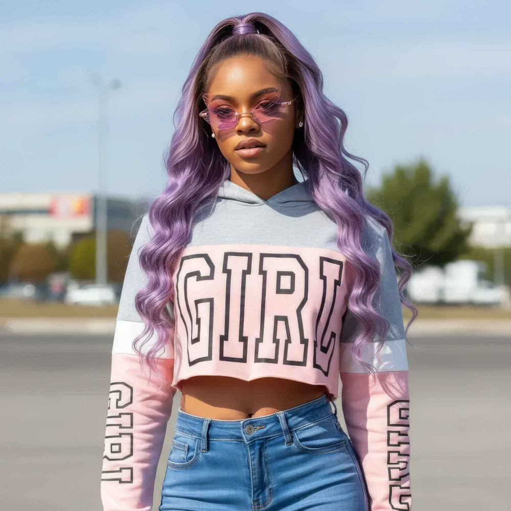 Woman with long vibrant purple wavy hair in a high ponytail wearing pink-tinted sunglasses and a color-block cropped hoodie featuring bold GIRL lettering across the chest with matching text on the sleeves. Styled with light blue denim jeans in an outdoor setting
