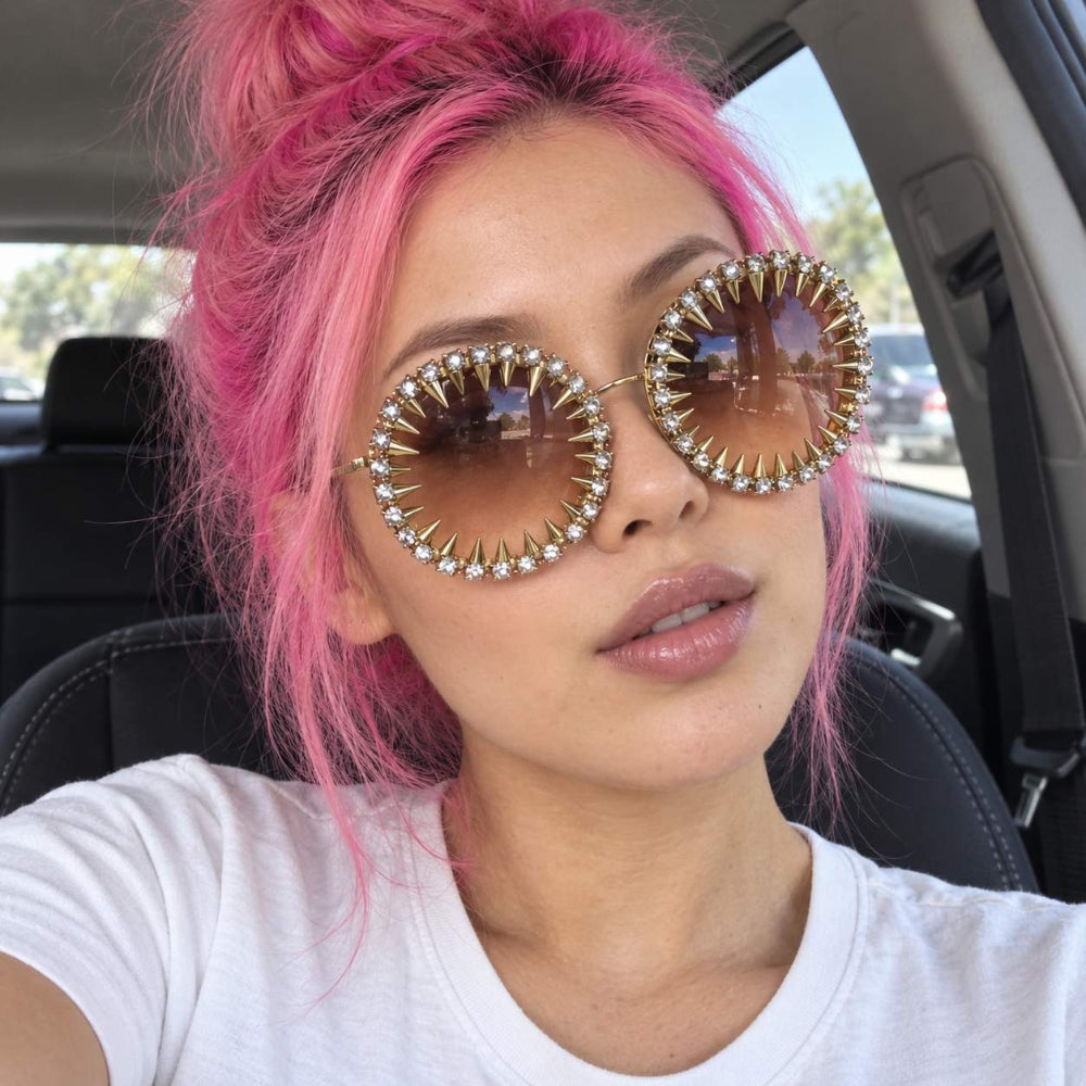 Woman with pink hair wearing gold round spike sunglasses with rhinestones and brown gradient lenses.