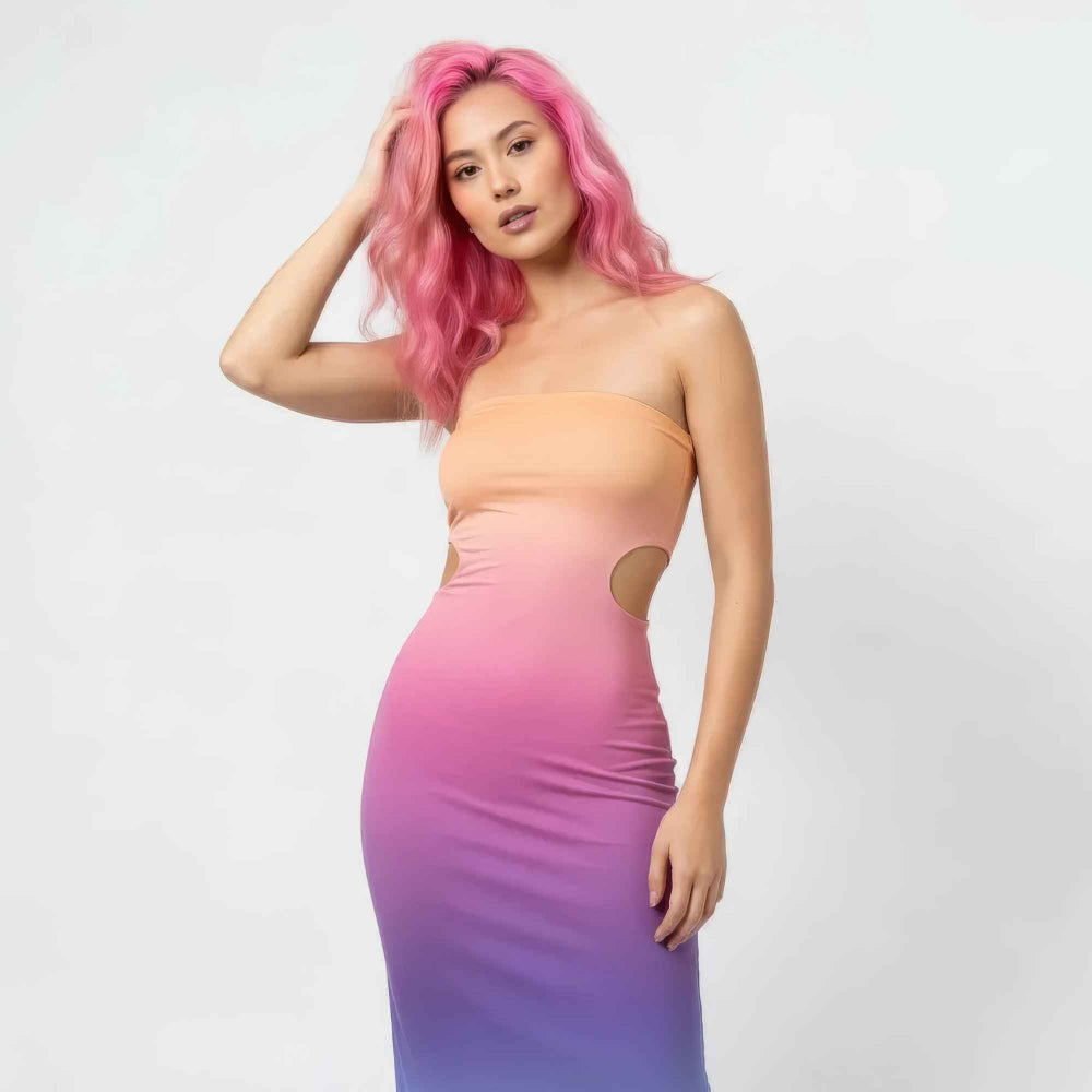 Front view of a woman with vibrant pink wavy hair modeling a strapless gradient ombre maxi dress transitioning from peach to pink to purple. The form-fitting dress features side cut-out details at the waist for a stylish, curve-hugging silhouette.