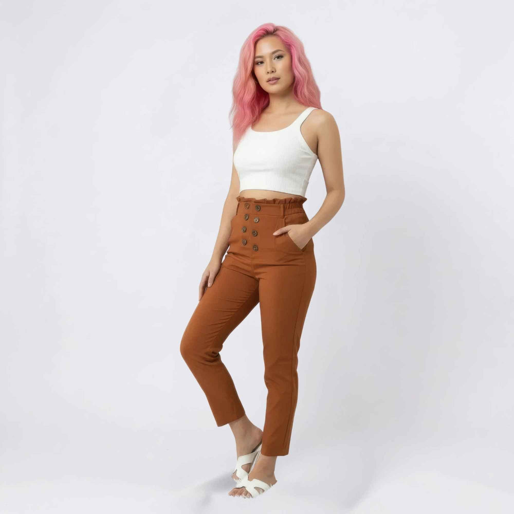 Full-length studio shot of a woman with vibrant pink hair modeling high-waisted brown trousers with a stylish double-row button detail at the waist and a paperbag waistband. She is wearing a white ribbed crop tank top, white strappy sandals, and poses with one hand in her pocket against a clean white background.
