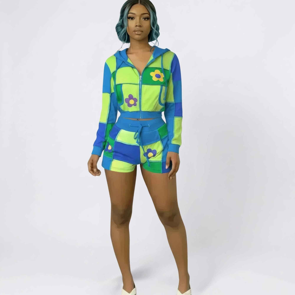 Full-length studio shot of a woman with teal wavy hair modeling a vibrant retro-style two-piece tracksuit. The set features a long-sleeve zip-up cropped hoodie and matching high-waisted shorts in a bold checkerboard pattern of lime green, blue, and purple, accented with colorful flower graphics and multiple pockets.