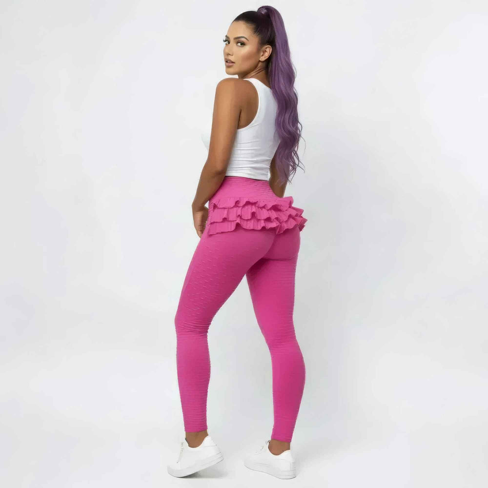 Back three-quarter view of a woman with long purple hair in a high ponytail modeling bright pink high-waisted leggings with dramatic multi-layered ruffle detailing around the hips. She is wearing a white cropped tank top and white sneakers, posing confidently to showcase the curve-enhancing fit.
