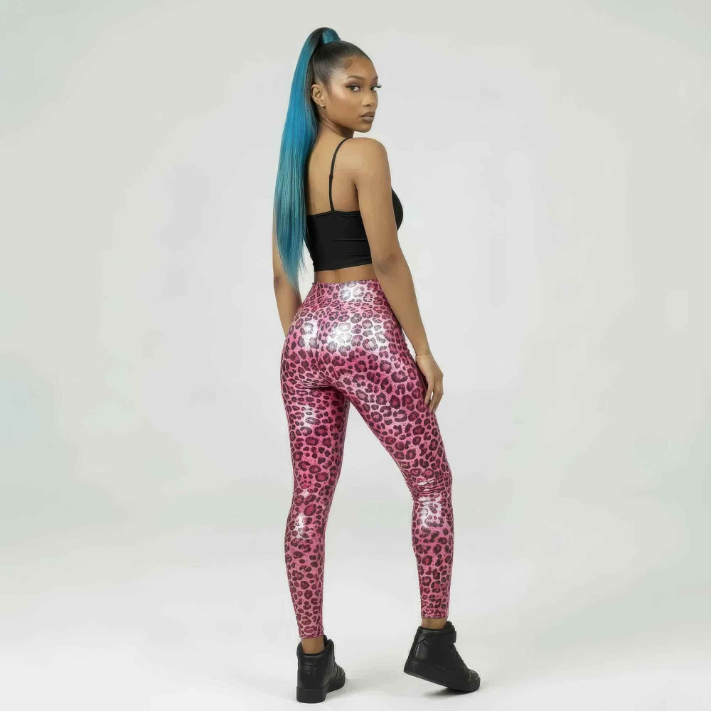 Back three-quarter view of a woman with long blue ponytail hair modeling shiny pink leopard-print high-waisted leggings. She is wearing a black strappy crop top and black ankle boots, posing confidently while looking over her shoulder to showcase the form-fitting, glossy animal print design.