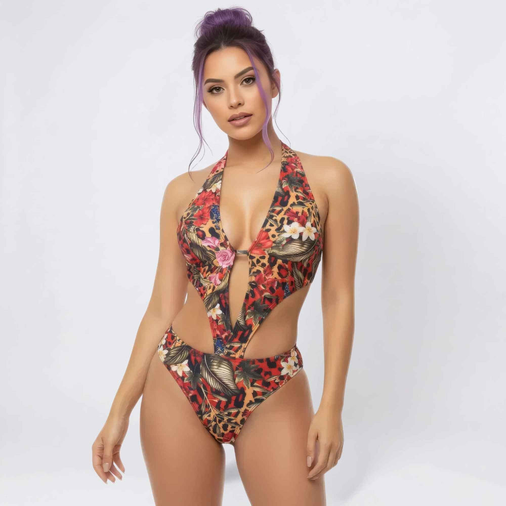Front view of a woman with purple hair in a high bun modeling a bold multicolored one-piece swimsuit featuring a deep plunging halter neckline with a central cut-out and intricate floral, animal print, and leaf patterns in red, black, and gold tones.