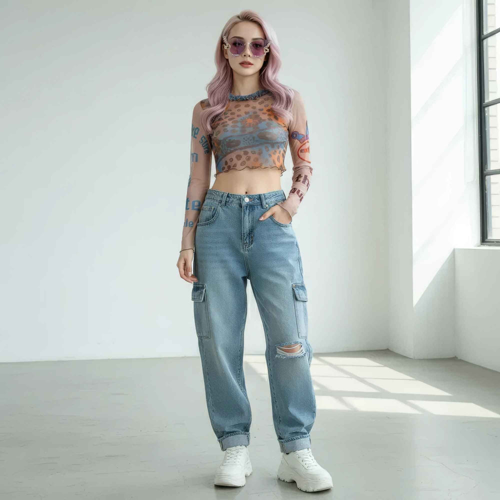 Woman with long pastel pink hair wearing ornate decorative sunglasses, a sheer long-sleeve mesh crop top with colorful leopard print, and light blue high-waisted distressed cargo jeans with knee rips. Styled with white chunky sneakers and visible arm tattoos, posing confidently in a bright studio setting.