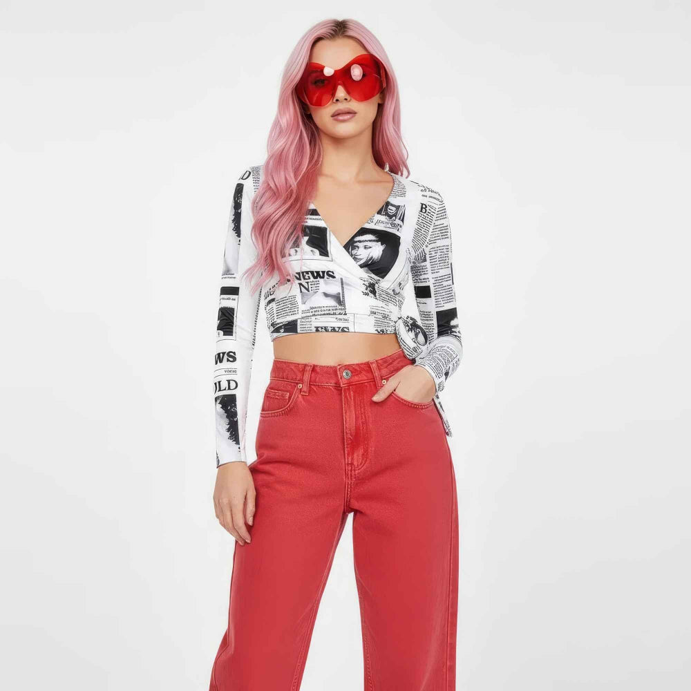 Woman with long vibrant pink wavy hair wearing oversized red heart-shaped sunglasses, a cropped long-sleeve newspaper print top, and high-waisted red wide-leg pants. She poses confidently with one hand in her pocket against a plain white background.