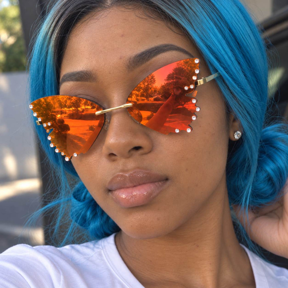 Woman with blue hair wearing orange rimless butterfly sunglasses with rhinestone edges.