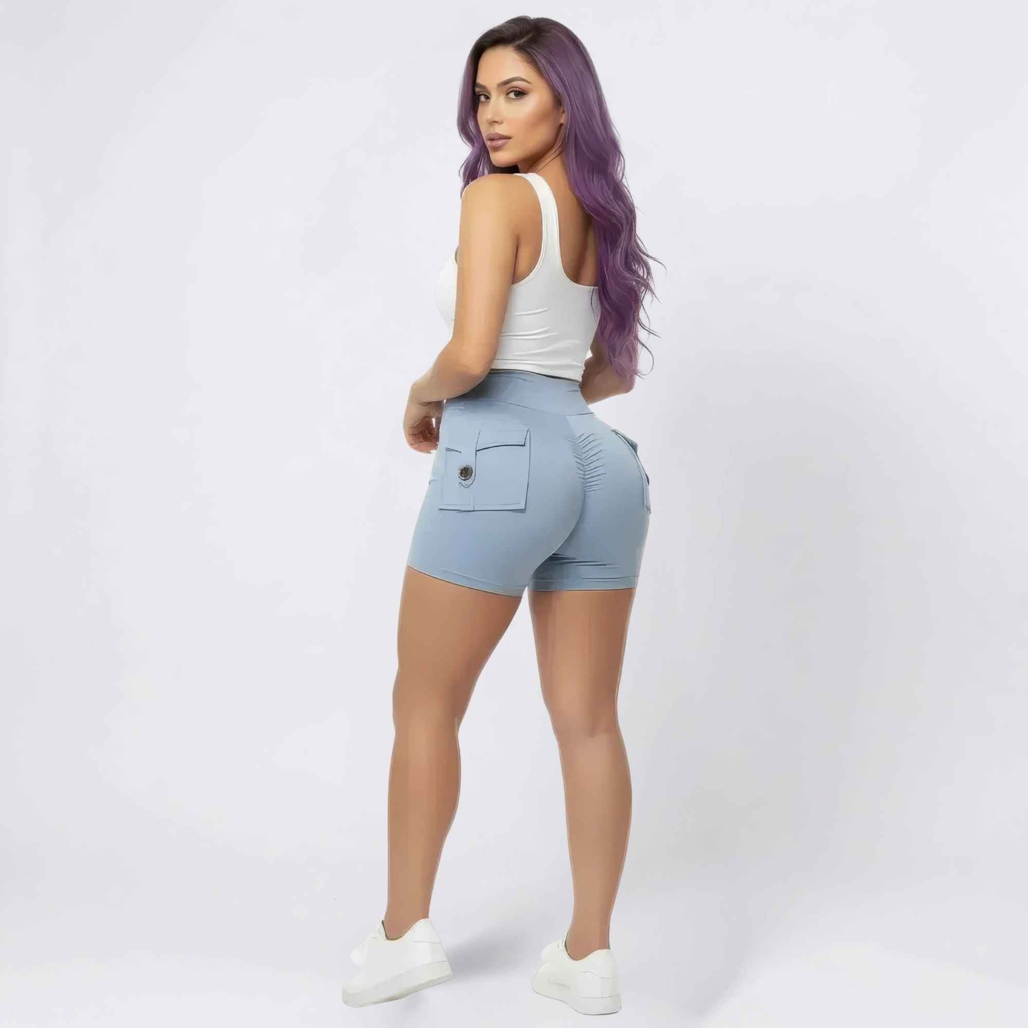 Back three-quarter view of a woman with long vibrant purple hair wearing a white sleeveless tank top and light blue high-waisted cargo shorts with back flap pockets and seam detailing. The shorts feature a sculpted, curve-hugging fit. Styled with white sneakers, posing confidently in a studio setting.