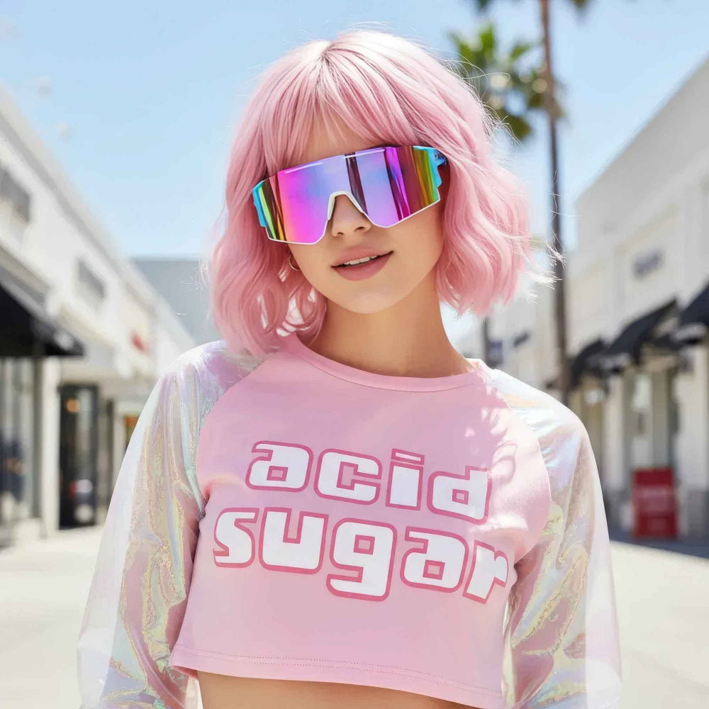 Woman with short vibrant pink bob hair and bangs wearing oversized iridescent visor sunglasses and a pink cropped t-shirt with bold acid sugar graphic text. The top features sheer holographic long sleeves, creating a playful Y2K-inspired street style look on a sunny urban street.