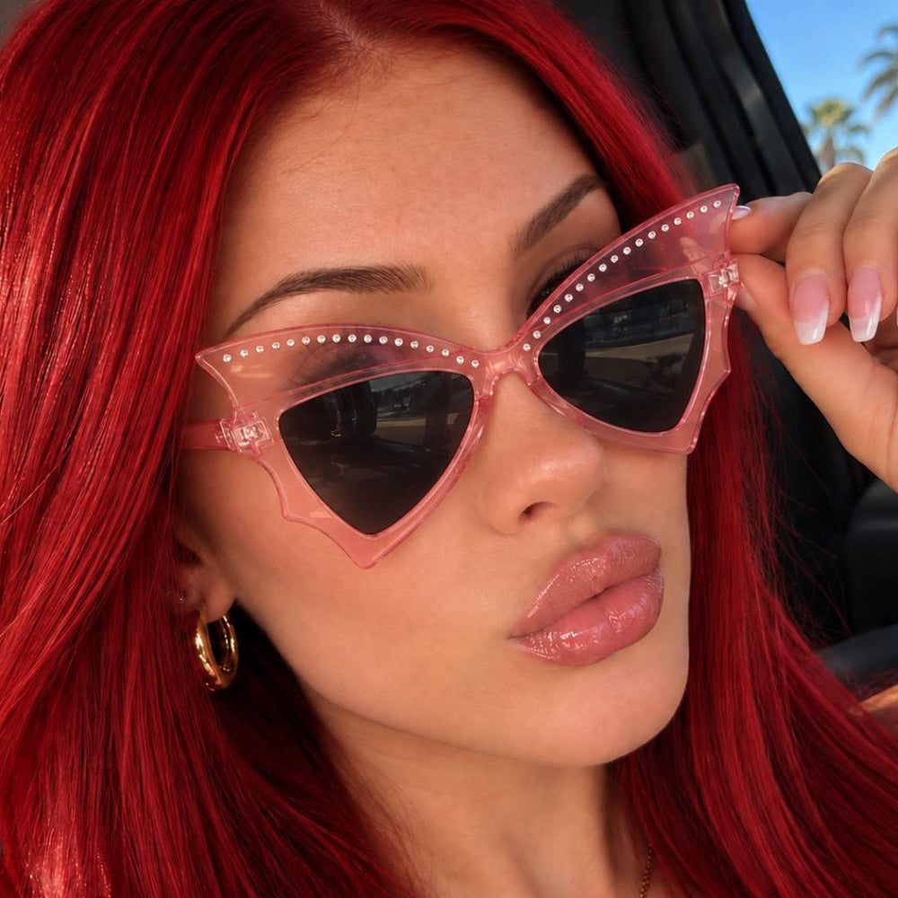 Woman with red hair wearing pink studded bat-wing sunglasses with dark lenses.