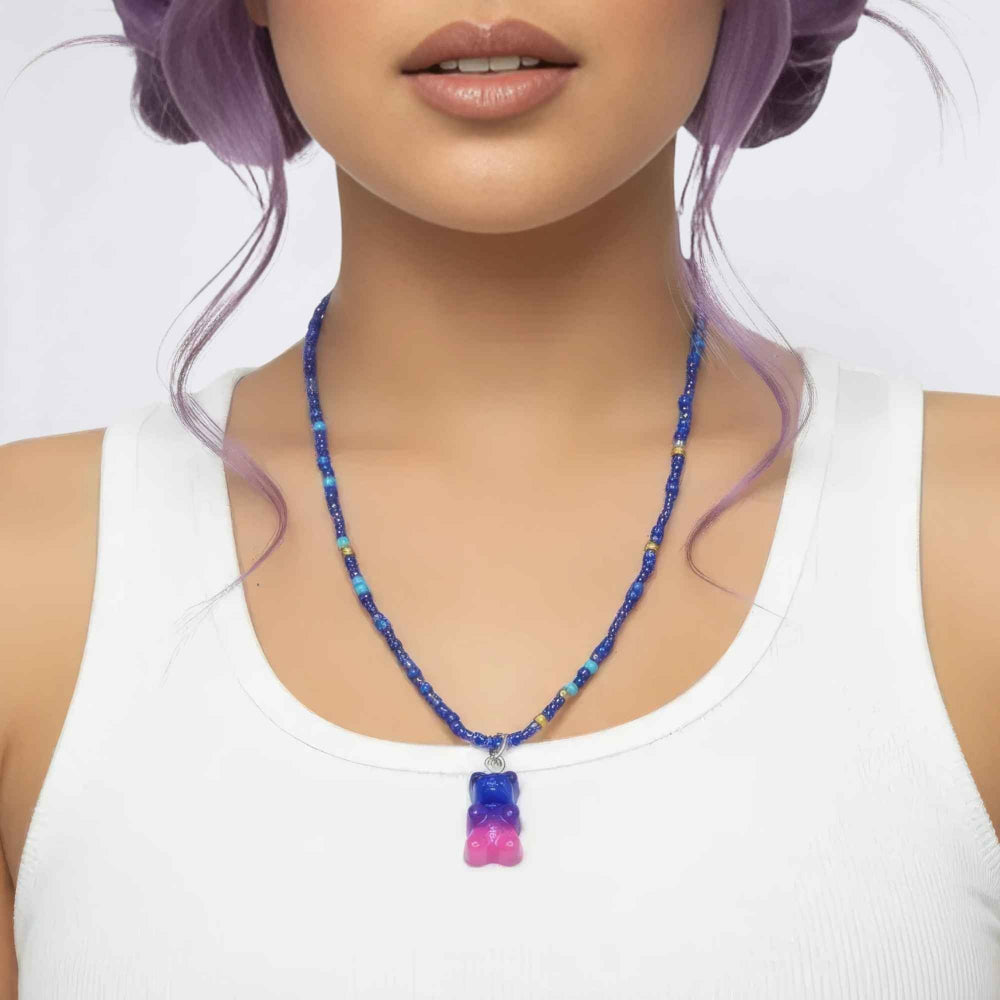 Close-up of a woman with lavender purple hair wearing a beaded necklace with blue, turquoise, and gold beads, featuring a gradient blue-to-pink gummy bear pendant, paired with a white tank top.