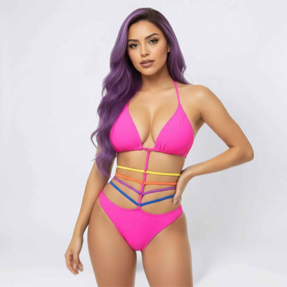 Front view of a woman with long vibrant purple wavy hair modeling a bright pink two-piece bikini. The set features a triangle halter top and high-cut bottoms with a bold cut-out waist design accented by colorful rainbow straps in yellow, orange, purple, and blue.