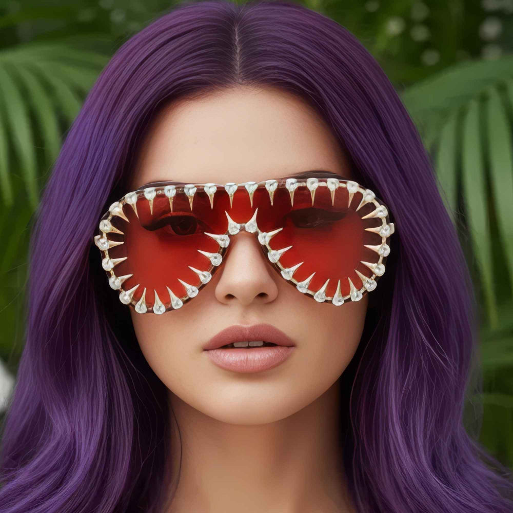 "Woman with vibrant purple hair wearing ornate steampunk sunglasses with red lenses and silver jeweled frame, against a green leafy background"