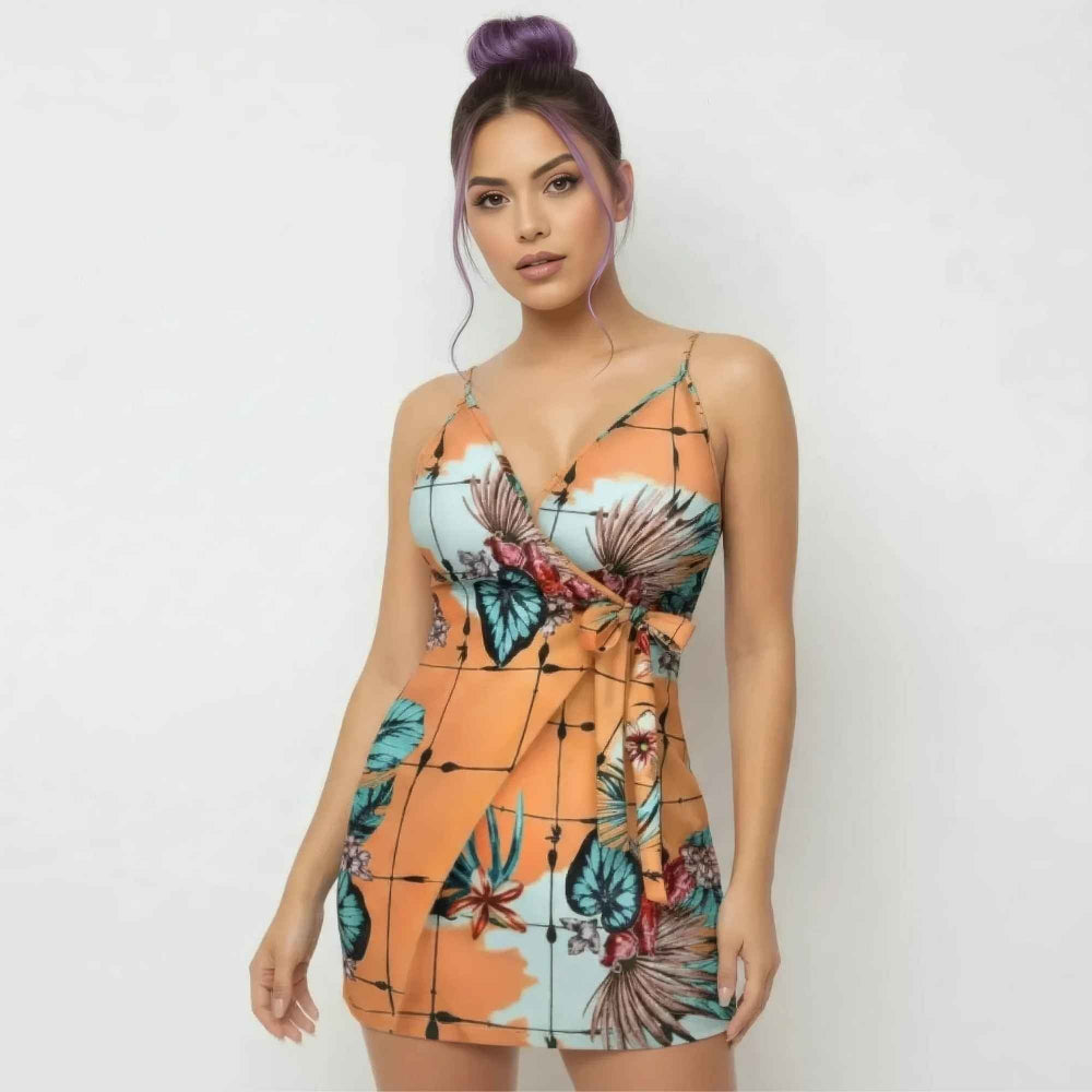 Front view of a woman with purple hair styled in a high bun modeling a vibrant tropical print mini dress. The form-fitting orange dress features thin spaghetti straps, a deep V-neckline, colorful floral and leaf patterns, and a stylish side tie wrap detail at the waist.