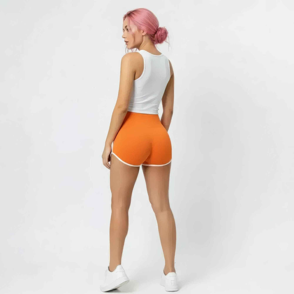 Back view of a woman with bright pink hair in a bun wearing a white sleeveless tank top and vibrant orange high-waisted booty shorts with white trim. The shorts feature a tight, sculpted fit that accentuates curves. Styled with white sneakers, posing in a studio setting.