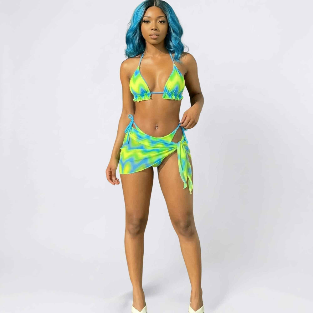 Full-length studio shot of a woman with vibrant blue wavy hair modeling a neon yellow and blue tie-dye bikini set. The look includes a triangle halter top with ruffled trim and matching bikini bottoms, paired with a coordinating tie-side sarong wrap skirt. She poses confidently with one hand touching the sarong.