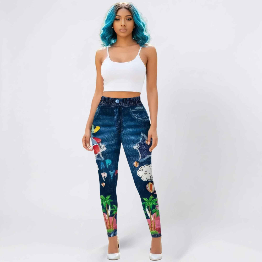Full-length front view of a woman with vibrant blue wavy hair modeling high-waisted dark blue graphic jeans featuring colorful tropical and summer prints including palm trees, beach balls, stars, clouds, and hot air balloons. She is wearing a white spaghetti strap crop top and white heels, posing confidently in a studio setting.