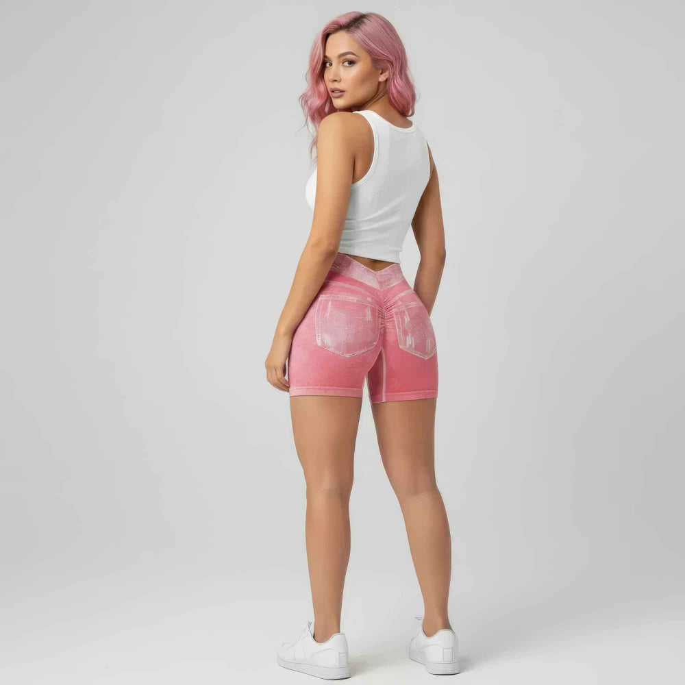 Back three-quarter view of a woman with vibrant pink hair wearing a white sleeveless crop top and light pink high-waisted distressed denim shorts with visible back pockets and faded details. The shorts feature a curve-hugging fit. Styled with white sneakers, posing confidently in a studio setting.