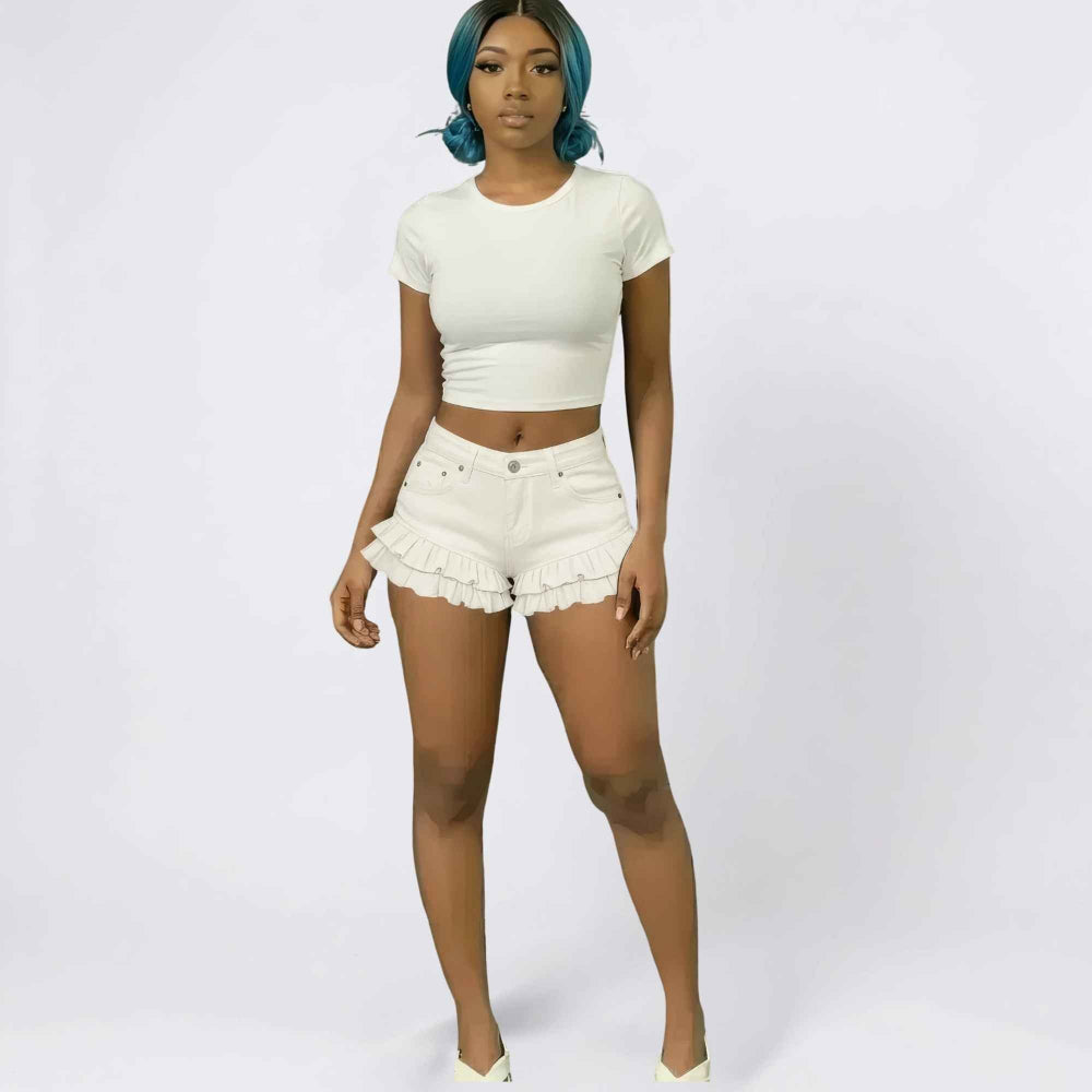 Woman with vibrant blue hair modeling a white short-sleeve crop top and white ruffled denim shorts with a flattering high-waisted fit. Full-body studio shot for Boutique Jolie fashion collection.