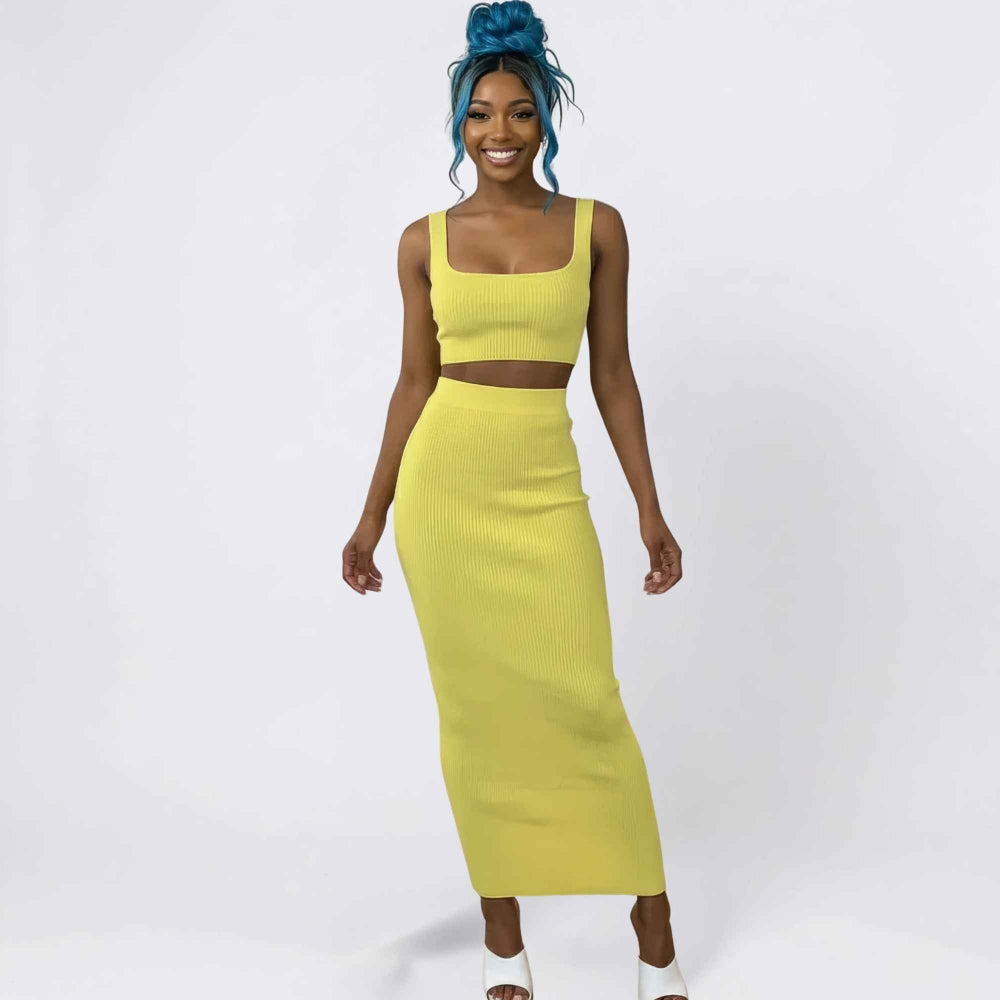 Full-length studio shot of a smiling woman with vibrant blue hair in a high messy bun modeling a bright yellow ribbed two-piece set. The outfit includes a square-neck crop top and a high-waisted, form-fitting maxi skirt, paired with white heeled sandals.
