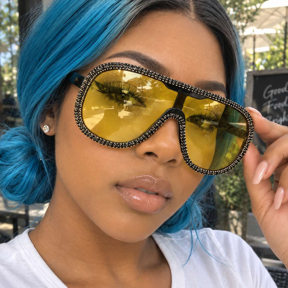Woman with blue hair wearing oversized yellow-lens sunglasses with a crystal-trimmed shield frame.