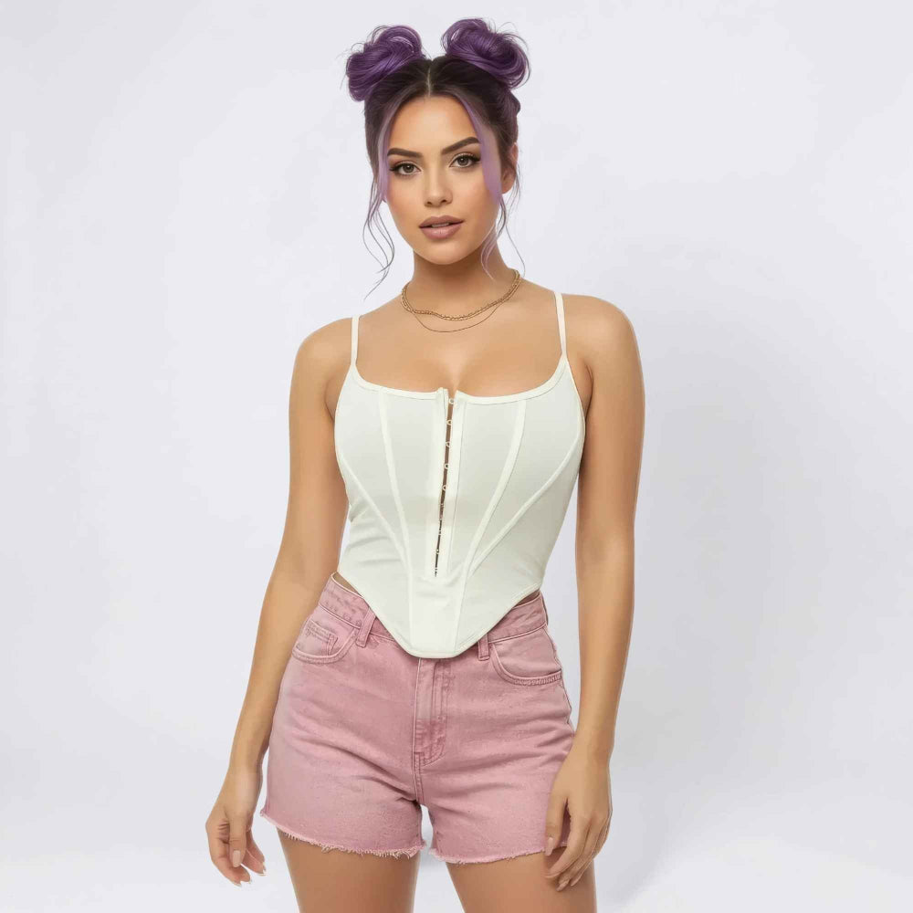 Front view of a woman with purple hair in playful space buns modeling a cream-colored structured corset bodysuit with front zipper detail and boning, paired with light pink high-waisted denim shorts. She wears layered gold necklaces and poses confidently against a plain white background.