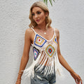 Blonde woman wearing a vibrant multicolored crochet crop top with granny square patterns, spaghetti straps, and long white fringe tassels. Styled with distressed blue denim jeans and gold jewelry, posing against a white textured wall.