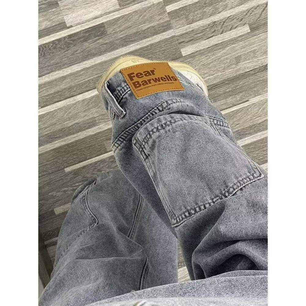 Close-up view of gray denim jeans with a visible brown "Fear Barwells" brand leather patch/label on the waistband, laid out or worn on a wooden floor.
