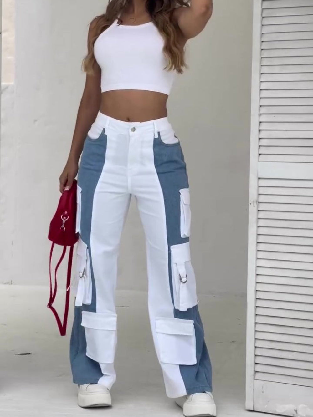 Woman with long wavy hair modeling a white crop top and color-blocked white and blue high-waisted cargo pants with multiple utility pockets, holding a red mini bag and styled with white sneakers. Full-body casual fashion shot for Boutique Jolie.