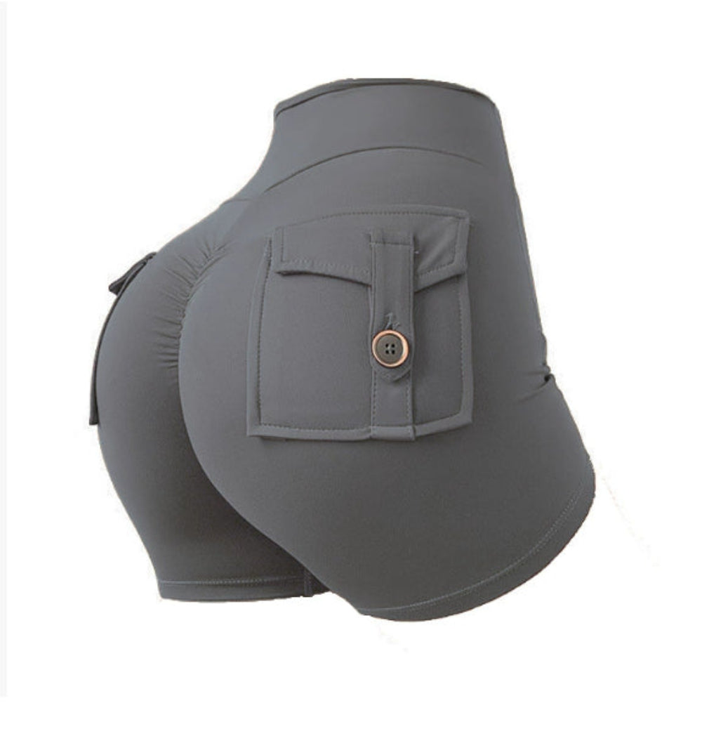 Woman modeling gray high-waisted athletic cargo shorts with a large back flap pocket and button detail, shown from a rear three-quarter view against a clean white studio background.