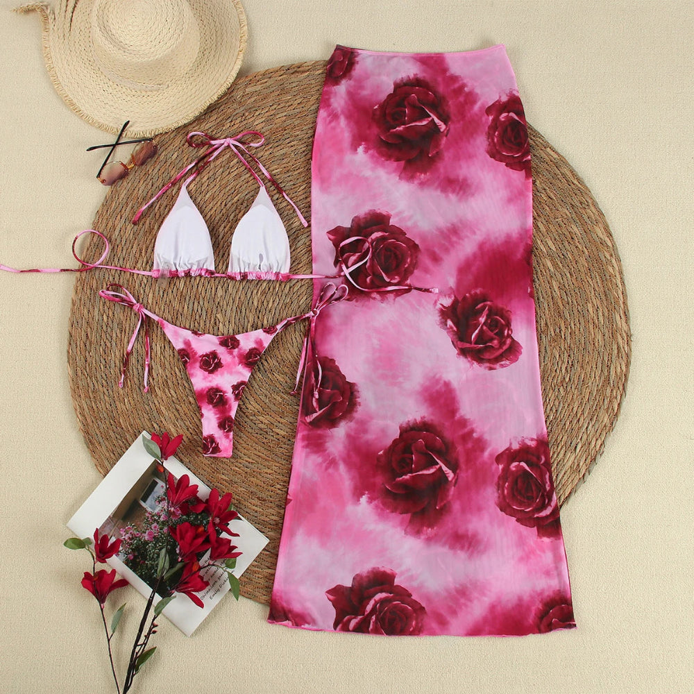 White bikini set with pink floral pattern on a pink floral towel, straw hat, and flowers on a beige background.