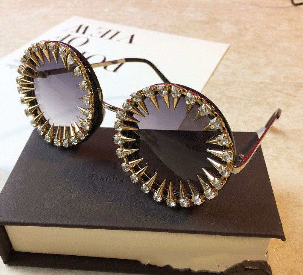 Black round steampunk sunglasses with gold spikes, rhinestones, and dark gradient lenses, displayed on a brown box.